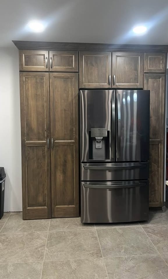 A floor-to-ceiling medium-brown stained pantry cabinet with a pair of raised-panel doors, beside a black built-in refrigerator