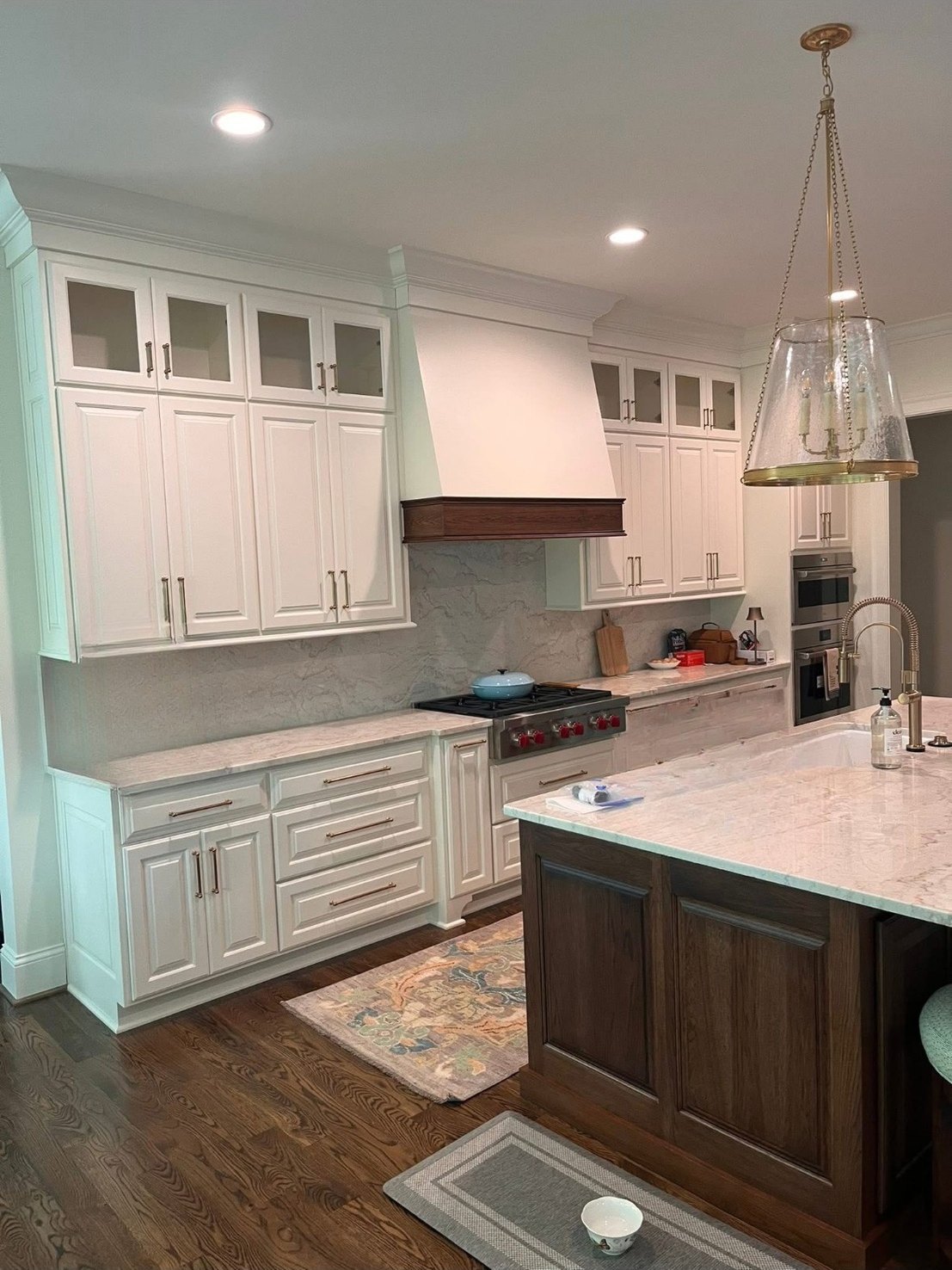 A white raised-panel kitchen with glass-front uppers, a white hood with a stained wood band, a dark-stained island and a glass pendant light