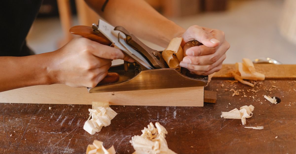 A cabinetmaker's hands using a hand plane to smooth a wooden board, shavings curling off the blade